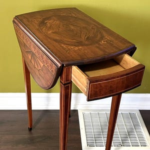 May include: A small, antique wooden side table with a drop-leaf design. The table features a dark brown finish with inlaid decorative patterns on the tabletop and drawer. The drawer is open, revealing the interior. The table stands on slender, tapered legs.