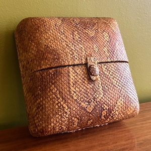 May include: A rectangular box with rounded corners, covered in a snakeskin pattern. The box is tan and brown with a small clasp closure. The box is sitting on a wooden surface with a green background.