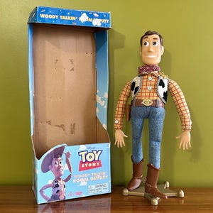 May include: A Woody action figure from the Disney Pixar Toy Story franchise stands next to its original box. The figure wears a yellow plaid shirt, blue jeans, a brown belt, and cowboy boots. The box is blue with the words "Woody Talkin' Room Deputy".