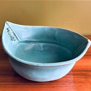 May include: A light blue ceramic bowl with a unique leaf-shaped design. The bowl has a smooth, glazed finish and a slightly rounded bottom.