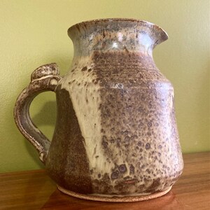 May include: A brown and white ceramic pitcher with a unique handle. The pitcher has a speckled glaze and a rounded body.