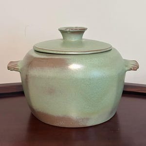 May include: A round ceramic pot with a lid, featuring a matte finish in shades of sage green and muted brown. The pot has two small, integrated side handles and a central knob on the lid. The surface shows subtle color variations and texture.