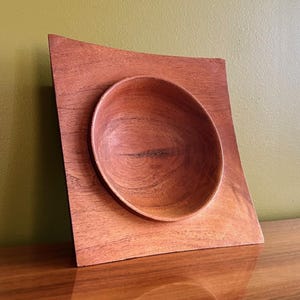 May include: A handcrafted wooden bowl with a square base and a rounded, recessed bowl. The wood has a warm reddish-brown hue, with visible grain patterns. The bowl is set on a wooden surface, showcasing a minimalist design.