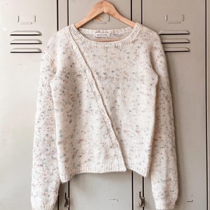 Op de afbeelding: Een wit gebreid vest met een gespikkeld patroon en een enkele knoopsluiting. Het vest hangt aan een houten hanger tegen een achtergrond van lockerdeuren.