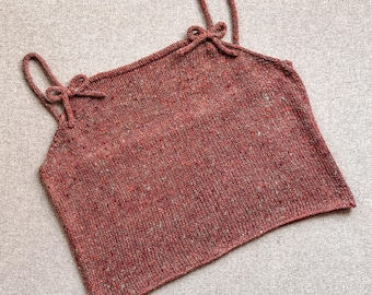 May include: A sleeveless knit camisole top in a speckled terracotta red yarn. The garment features a straight neckline and two thin shoulder straps, each accented with a small, decorative hand-tied bow at the point of attachment.
