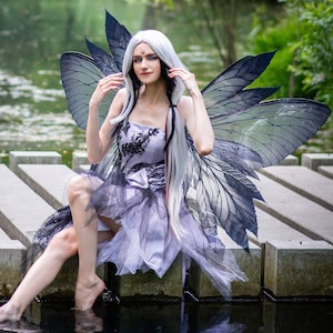 Puede incluir: Una mujer con el pelo largo y blanco y un disfraz de mariposa blanco y negro se sienta en un muelle de madera sobre un estanque. Lleva un vestido morado con encaje negro y un disfraz de mariposa blanco y negro.