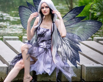 May include: A woman with long white hair and a black and white butterfly costume sits on a wooden dock over a pond. She is wearing a purple dress with black lace and a black and white butterfly costume.