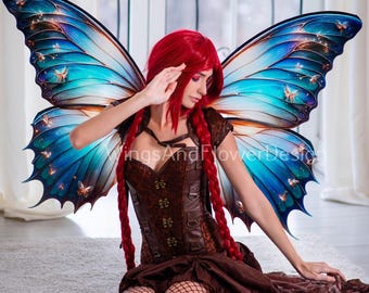 May include: A woman with long red hair wearing a brown corset and a brown skirt with a black fishnet stocking. She is wearing large blue and brown butterfly wings with gold glitter accents. The wings are spread out behind her.