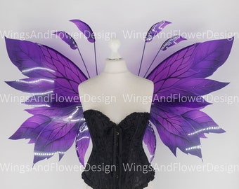 May include: A black corset with a floral pattern is displayed on a mannequin, paired with large, purple fairy wings. The wings have a detailed leaf design and are accented with white lights. The background is plain white.