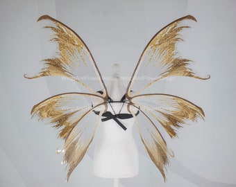 May include: A pair of fairy wings with a gold glitter design. The wings are attached to a black harness with a bow. The wings are displayed on a white mannequin. The text "WingsAndFlower Design" is printed on the wings.