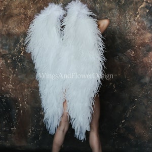 Puede incluir: Un par de alas de ángel grandes de plumas blancas. Las alas están hechas de plumas suaves y esponjosas y están unidas a la espalda de una persona. La persona está de pie frente a una pared marrón texturizada. El texto "WingsAndFlowerDesign" es visible en las alas.