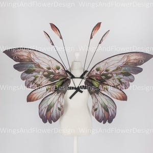 May include: A pair of large, intricately designed butterfly wings with a floral pattern in shades of brown, green, and purple. The wings are attached to a black harness and are displayed on a white mannequin.
