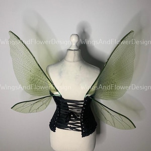 May include: A black corset with a lace-up back and attached green and translucent fairy wings. The wings have a delicate, veined pattern. The corset is displayed on a mannequin against a plain background.