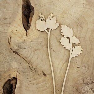 May include: Two wooden plant silhouettes with intricate leaf details. The plants are cut from a light brown wood and are placed on a light brown wood background.