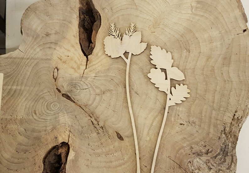May include: Two wooden plant silhouettes with intricate leaf details. The plants are cut from a light brown wood and are placed on a light brown wood background.