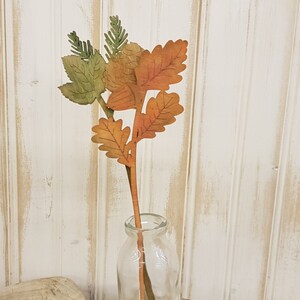 May include: A single stem of faux fall leaves in shades of orange, brown, and green. The leaves are arranged in a cluster at the top of the stem, and the stem is inserted into a clear glass vase.