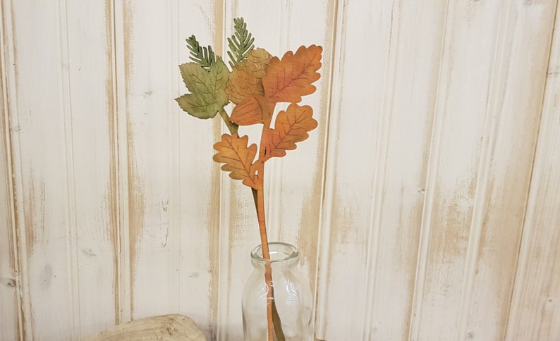 May include: A single stem of faux fall leaves in shades of orange, brown, and green. The leaves are arranged in a cluster at the top of the stem, and the stem is inserted into a clear glass vase.