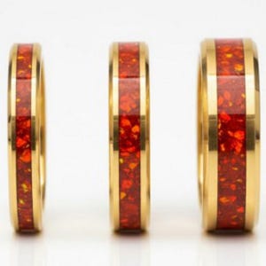 Op de afbeelding: Drie goudkleurige ringen met een levendige rood-oranje gespikkelde inleg. De ringen worden gepresenteerd tegen een witte achtergrond, die hun gepolijste afwerking en ingewikkelde ontwerp benadrukt. De ringen zijn waarschijnlijk voor een bruiloft of verloving.