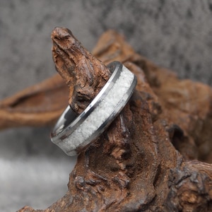 May include: A silver ring with a white stone inlay. The ring is set against a textured, brown wooden background. The ring's band is a polished silver color, and the inlay appears to be a crushed stone or composite material.