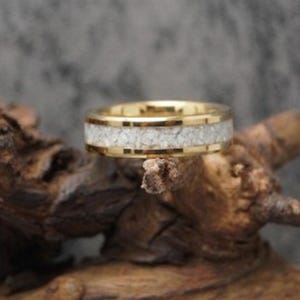 May include: A gold-colored ring with a white, textured inlay. The ring is set against a natural, wooden background. The band has a polished finish, and the inlay appears to be made of small, irregular pieces.