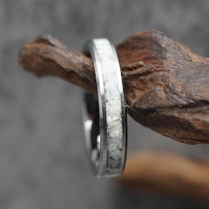 Puede incluir: Primer plano de un anillo plateado con una incrustación blanca texturizada. El anillo se encuentra sobre un fondo borroso y descansa sobre un trozo de madera marrón oscuro. El diseño del anillo es simple y elegante.