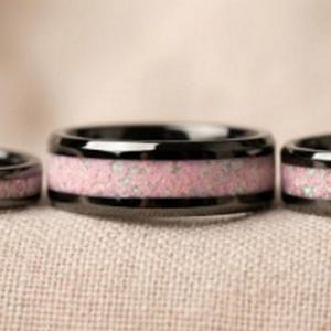 May include: Three black rings with a pink, speckled center band. The rings are displayed on a neutral-colored surface. The rings have a glossy finish.