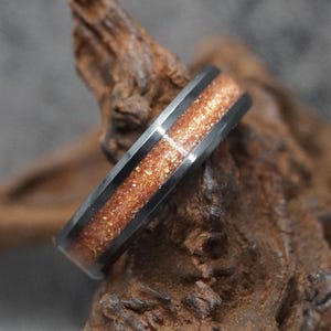 May include: A black ring with a copper-colored, glittery inlay. The ring is set against a textured, brown wooden background. The ring's band is a dark, metallic color.