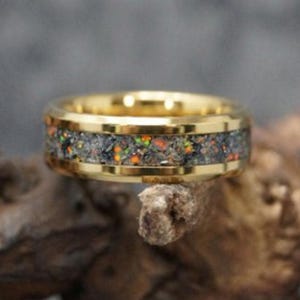 May include: A gold-colored ring with a band of dark gray, orange, and green speckled material. The ring is set on a piece of wood, with a blurred background.