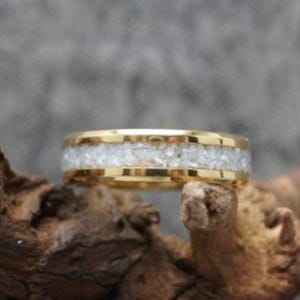 Puede incluir: Un anillo dorado con una banda central de material blanco texturizado. El anillo se muestra sobre un fondo borroso y descansa sobre un trozo de madera, resaltando su diseño y detalles.