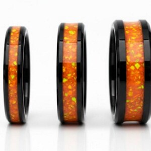 Könnte beinhalten: Drei schwarze Ringe mit einem lebendigen, orange-gelb gesprenkelten Inlay. Die Ringe werden auf einer weißen Oberfläche präsentiert, die ihre polierte Oberfläche und die kontrastierenden Farben zeigt. Es handelt sich wahrscheinlich um Schmuck.