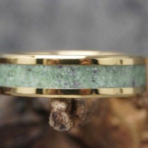 May include: A gold ring featuring a central band of speckled green stone. The ring has a polished finish, and the stone band is framed by the gold metal. The ring is a piece of jewellery.