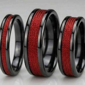 May include: Three black rings with a red woven center band. The rings have a polished black finish and a textured red center. The rings are likely for a couple or family.