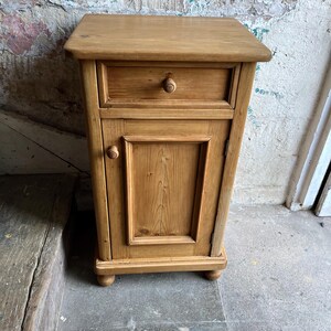 May include: A light brown wooden bedside table featuring a rectangular top, one upper drawer with a small round knob, and a lower cabinet door with a matching round knob. The piece is supported by four small, rounded feet at the base.