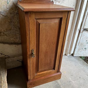 May include: A small, antique wooden cabinet with a single door. The cabinet is made of a warm-toned wood and features a decorative top and a brass-coloured handle. The cabinet is standing on a concrete surface.