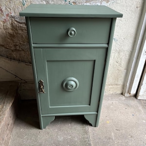 May include: A sage green bedside cabinet with a single drawer and a door. The cabinet has a round knob on the drawer and a larger, decorative circular element on the door. It features a small brass-colored handle and sits on decorative feet.