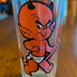 Puede incluir: Un vaso transparente con una ilustración de diablo de dibujos animados en rojo, negro y blanco. El diablo tiene cuernos, una cola y un tridente. Las palabras "HOT STUFF" están impresas en la parte inferior del vaso.