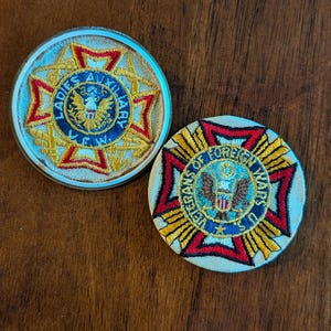 Może przedstawiać: Dwie okrągłe, haftowane naszywki. Jedna z napisem "Ladies Auxiliary" z emblematem orła. Druga z napisem "Veterans of Foreign Wars" z emblematem orła. Obie mają czerwone, złote i niebieskie akcenty.