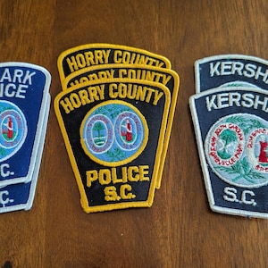 Puede incluir: Un conjunto de parches de policía bordados. Los parches presentan textos como "DENMARK POLICE S.C.", "HORRY COUNTY POLICE S.C.", y "KERSHAW S.C.". Los parches son de varios colores, incluyendo azul, negro, amarillo y blanco.