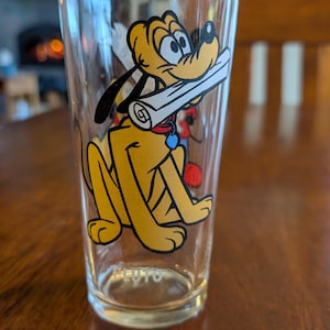 May include: Clear glass tumbler featuring a yellow cartoon dog, Pluto, holding a scroll in his mouth. The tumbler has a Pluto graphic on the front and the name "Pluto" at the bottom. The glass is on a wooden surface.
