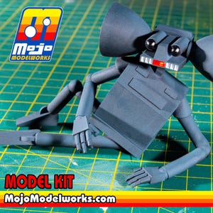 May include: A gray articulated robot model kit featuring large cone-shaped ears, two black spherical eyes, and a red mouth detail. The figure is in a reclining pose. The image includes the Mojo Modelworks logo and the text MODEL KIT MojoModelworks.com.