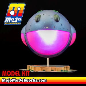 May include: A spherical model kit featuring a grey, metallic-textured surface with two dark oval eyes and a large, glowing pink circular mouth. The model is mounted on a thin silver post above a detailed tan building base. Text reads Mojo Modelworks.