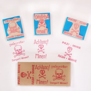 Puede incluir: Una colección de seis sellos de goma con un diseño de calavera y huesos cruzados. Los sellos están en diferentes idiomas, incluyendo inglés, alemán, ruso y tailandés. Los sellos están sobre un fondo blanco.