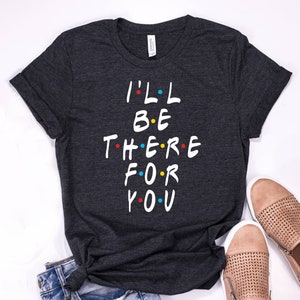 以下が含まれることがあります： 「I'LL BE THERE FOR YOU」の文字が白で書かれた濃いグレーのTシャツ。カラフルなドットが施されています。シャツは、ベージュのスリッポンシューズとデニムジーンズと組み合わせています。このシャツはカジュアルウェアです。