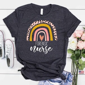 Puede incluir: Una camiseta gris oscuro con un dise&ntilde;o de arco&iacute;ris con rayas rosas, amarillas y naranjas. El arco&iacute;ris tiene un estampado de leopardo y un coraz&oacute;n rosa en el centro. El texto "FORENSIC nurse" est&aacute; escrito debajo del arco&iacute;ris.