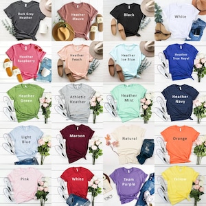 May include: A collage of 20 different coloured t-shirts, each with a different colour and text description. The t-shirts are arranged in a grid of four rows and five columns. The colours include dark grey heather, heather mauve, black, white, heather raspberry, heather peach, heather ice blue, heather true royal, heather green, athletic heather, heather mint, heather navy, light blue, maroon, natural, orange, pink, white, team purple, and yellow.
