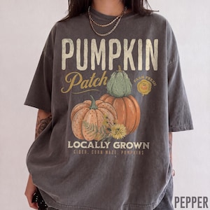 May include: A charcoal gray short-sleeve t-shirt featuring a graphic print with the text PUMPKIN PATCH, FARM FRESH, and LOCALLY GROWN. The design includes illustrations of orange and green pumpkins, yellow flowers, and the words CIDER, CORN MAZE, PUMPKINS.