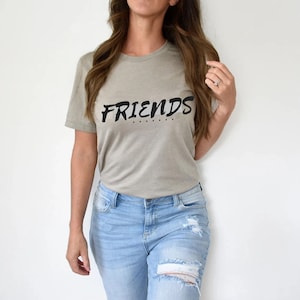 Puede incluir: Una camiseta gris claro con la palabra "FRIENDS" impresa en negro. La camiseta tiene un corte casual y mangas cortas.