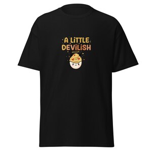Könnte beinhalten: Schwarzes T-Shirt mit dem Text "A Little Devilish" in Gold und Orange. Die Grafik zeigt einen Cartoon-Cupcake mit Heiligenschein und Herzdetails. Das Shirt ist ein kurzärmeliger, lässiger Stil.