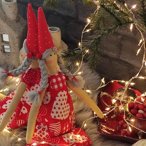May include: A handmade doll wearing a red dress with white hearts and a red hat with white polka dots. The doll has gray braids and is sitting on a fluffy gray rug with white Christmas lights.