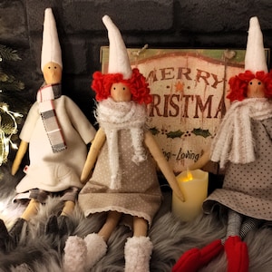 May include: Three handmade Christmas dolls with white fabric hats and red yarn hair. The dolls are sitting on a fluffy grey surface with a sign that reads "Merry Christmas" in the background.